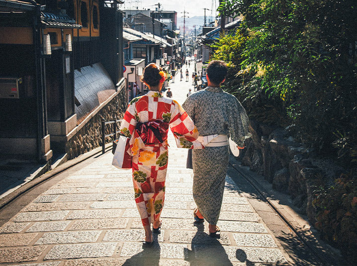 Two women in colorful kimono walking down a sunlit stone-paved street lined with traditional wooden machiya townhouses in Kyoto