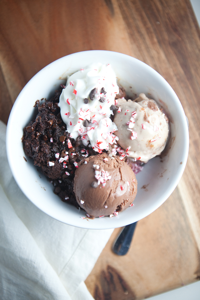 A bowl with two scoops of chocolate protein muffin ice cream, a brownie, whipped cream, chocolate chips, and crushed peppermint on top, sitting on a wooden surface with a white napkin underneath.