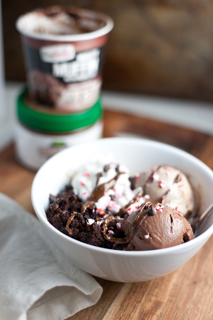 A white bowl with scoops of chocolate and vanilla ice cream, brownie pieces, whipped cream, chocolate drizzle, and crushed peppermint. The dessert is inspired by the rich flavors of chocolate protein muffin ice cream. Ice cream containers are blurred in the background on a wooden surface.