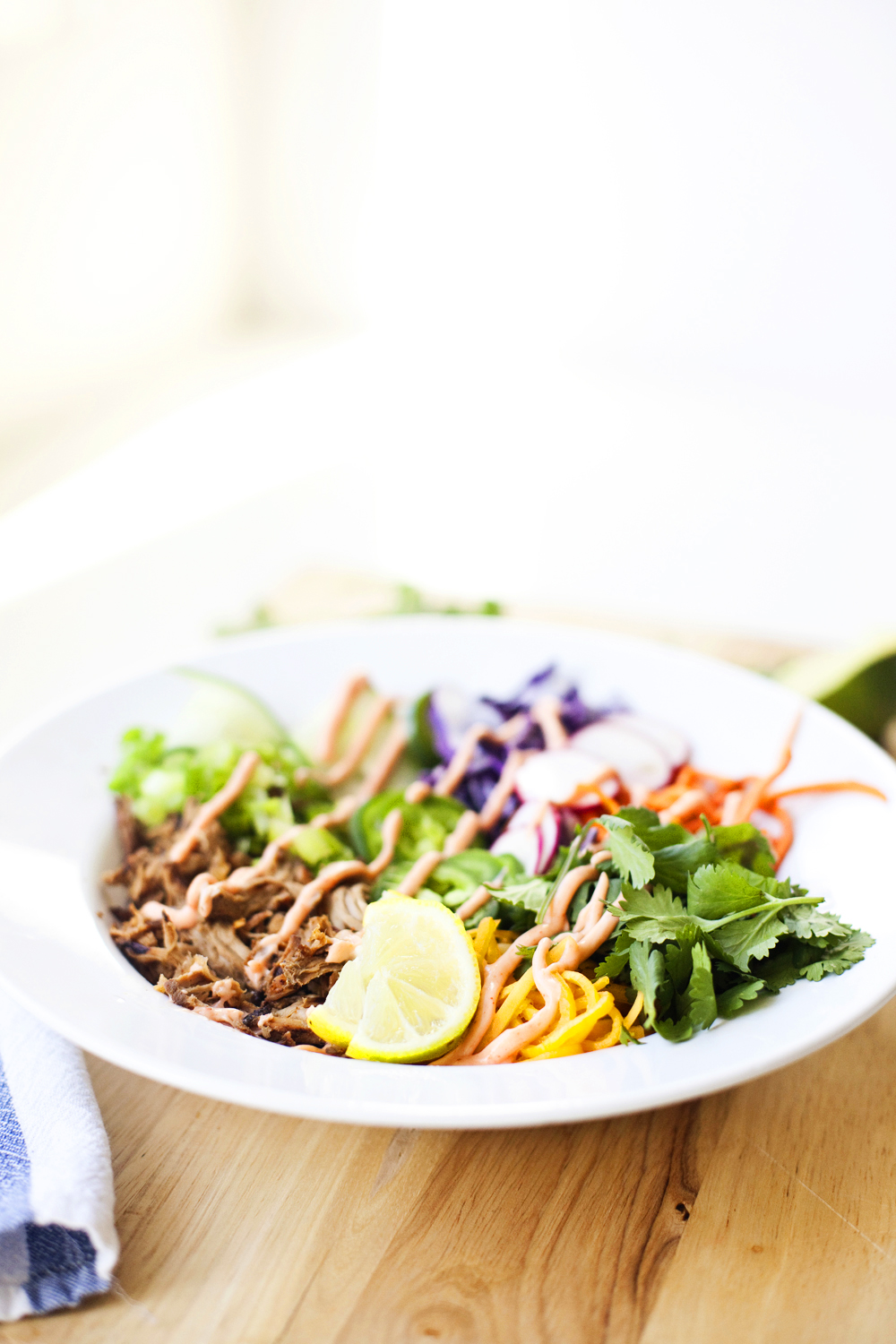 A white bowl filled with shredded meat, sliced vegetables, cilantro, and lime wedges sits on a wooden surface, bright natural light in the background—perfect for a quick lunch or as a fresh twist on a Banh Mi-inspired veggie bowl.