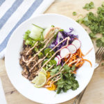 A vibrant Veggie Bowl featuring shredded meat, spiralized carrots, purple cabbage, radish slices, cucumber, lime, cilantro, and a drizzle of dressing sits on a white plate beside a fork and striped napkin—perfect for a quick lunch or your next Lunch-Time Banh Mi-inspired meal.