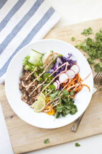 A vibrant Veggie Bowl featuring shredded meat, spiralized carrots, purple cabbage, radish slices, cucumber, lime, cilantro, and a drizzle of dressing sits on a white plate beside a fork and striped napkin—perfect for a quick lunch or your next Lunch-Time Banh Mi-inspired meal.