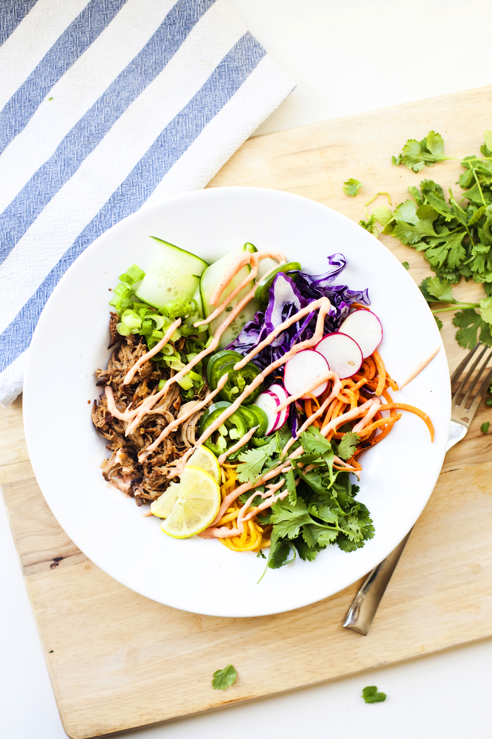 A vibrant Veggie Bowl perfect for Lunch-Time: A white bowl filled with shredded meat, spiralized carrots, purple cabbage, cucumber, radish slices, cilantro, and lime, drizzled with sauce in true Banh Mi style, sits on a wooden board beside a fork and a striped napkin.