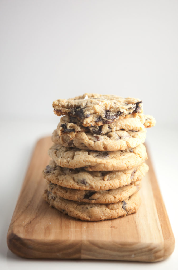 Stack of giant chocolate chip cookies on wood board with cookie on top broken in half.