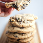 Stack of chocolate chip cookies on wood board with hand holding one cookie broken in half.