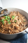 Shredded seasoned meat garnished with fresh cilantro in a large pan, with metal tongs resting on the edge, sitting on a dark blue cloth—these Easy Low-Fat Slow Cooker Crispy Carnitas offer a flavorful and healthier twist on a classic favorite.