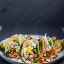 Three grilled fish tacos topped with shredded cabbage, mango salsa, avocado slices, and chopped herbs are served on a white plate with lime wedges on the sides—perfect alongside flavorful Chipotle Lime Instant Pot Taco Chicken for a vibrant fiesta spread.