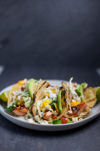 Three grilled fish tacos topped with shredded cabbage, mango salsa, avocado slices, and chopped herbs are served on a white plate with lime wedges on the sides—perfect alongside flavorful Chipotle Lime Instant Pot Taco Chicken for a vibrant fiesta spread.