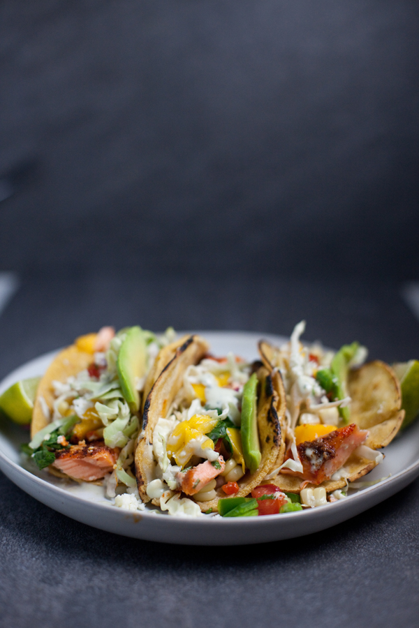 Three grilled fish tacos topped with shredded cabbage, mango salsa, avocado slices, and chopped herbs are served on a white plate with lime wedges on the sides—perfect alongside flavorful Chipotle Lime Instant Pot Taco Chicken for a vibrant fiesta spread.