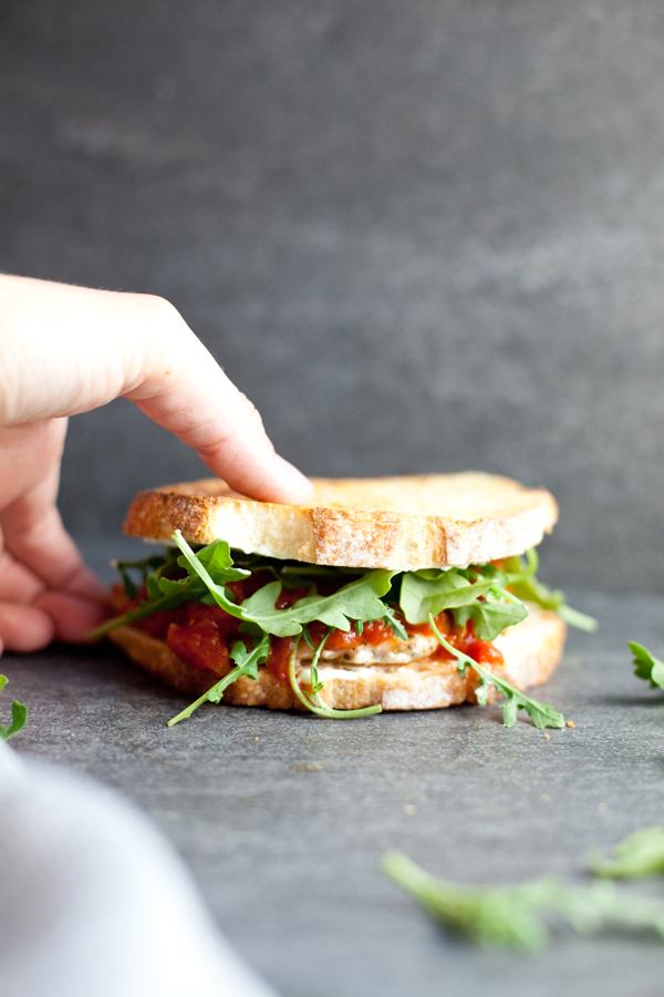A hand holds a sandwich filled with greens, tomato sauce, and savory Chipotle Lime Instant Pot Taco Chicken, on a gray surface with some scattered arugula leaves.