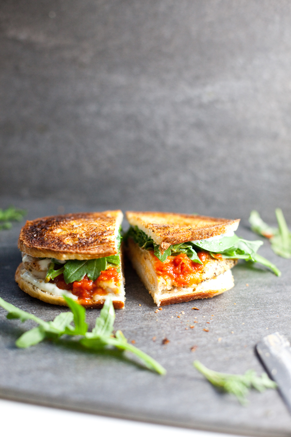 A grilled sandwich cut in half sits on a dark surface, filled with tomato sauce, melted cheese, fresh arugula, and tender Chipotle Lime Instant Pot Taco Chicken. Some arugula leaves are scattered around the sandwich.