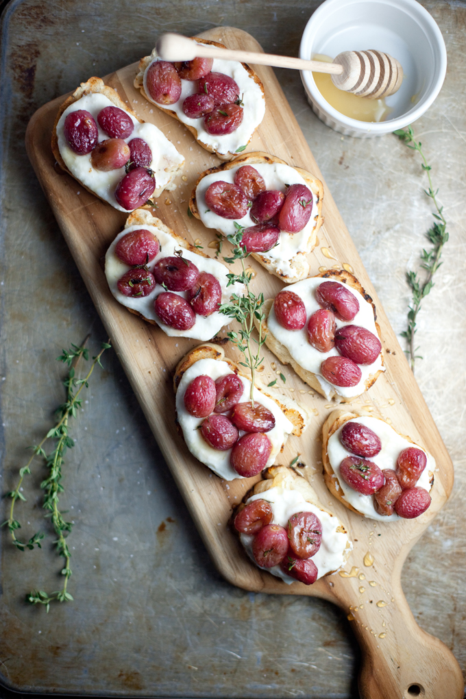 Toasted bread slices topped with melted cheese and roasted grapes, garnished with fresh thyme, on a wooden board—an elegant addition to any Thanksgiving menu. A honey dipper rests in a small bowl of honey nearby.