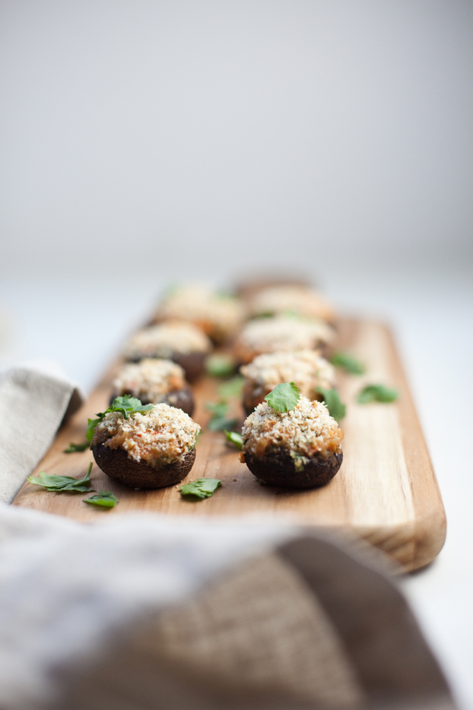 Stuffed mushrooms topped with breadcrumbs and chopped herbs are elegantly arranged on a wooden serving board, making them a perfect addition to your Thanksgiving menu. A neutral napkin sits in the foreground, while the background remains softly blurred for a cozy holiday touch.