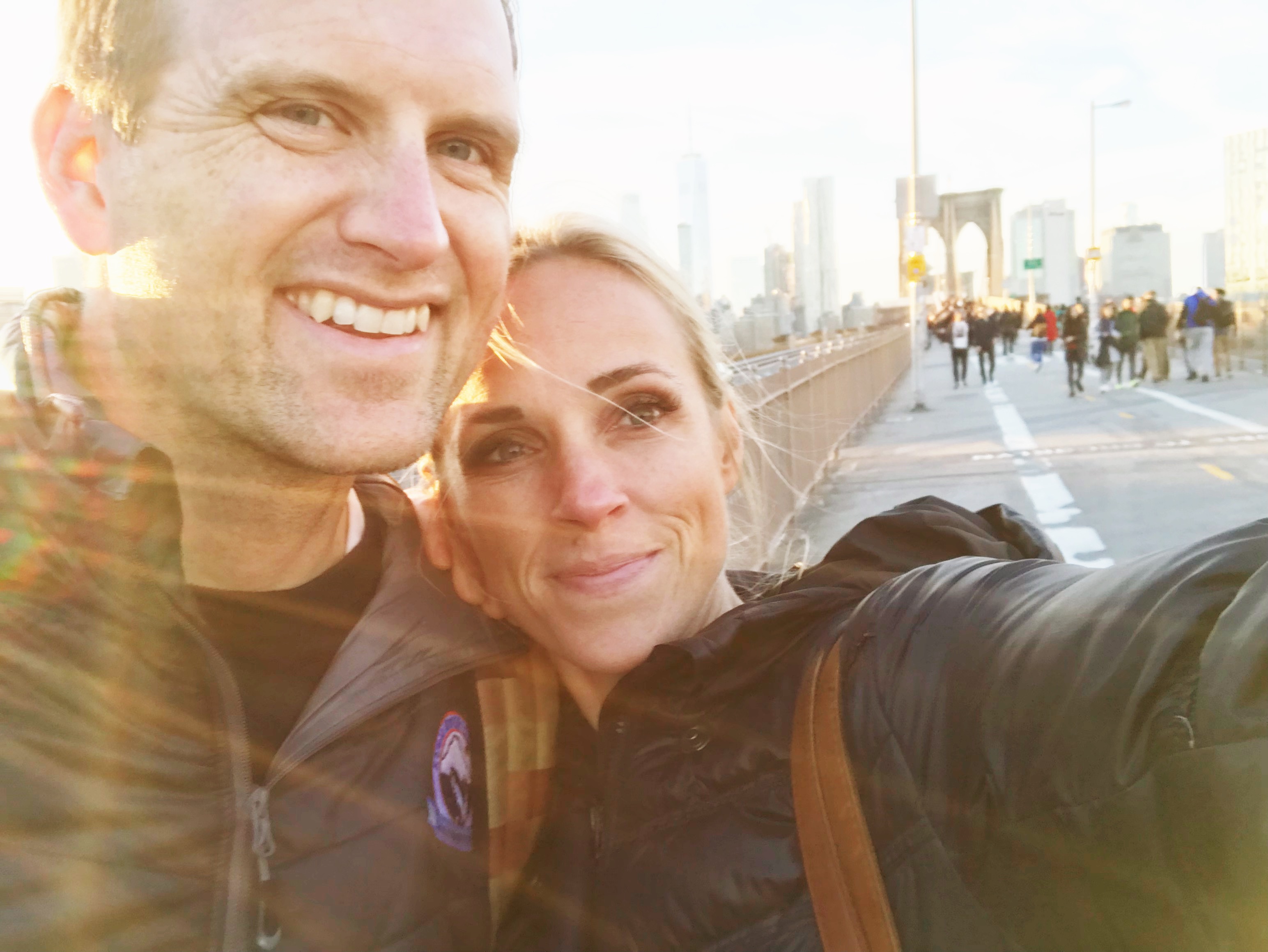 A smiling couple takes a selfie on a city bridge in daylight, surrounded by pedestrians. Sunlight creates a warm glow and lens flare, with tall buildings visible in the background—perhaps they’re celebrating their success with macro counting as they capture this vibrant moment together.