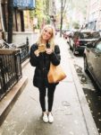 A woman in a black jacket stands on a city sidewalk, smiling and holding a large cookie—perhaps thinking about macro counting as she enjoys her treat. She has a tan bag over her shoulder and wears black pants and white shoes. Cars and trees line the street.