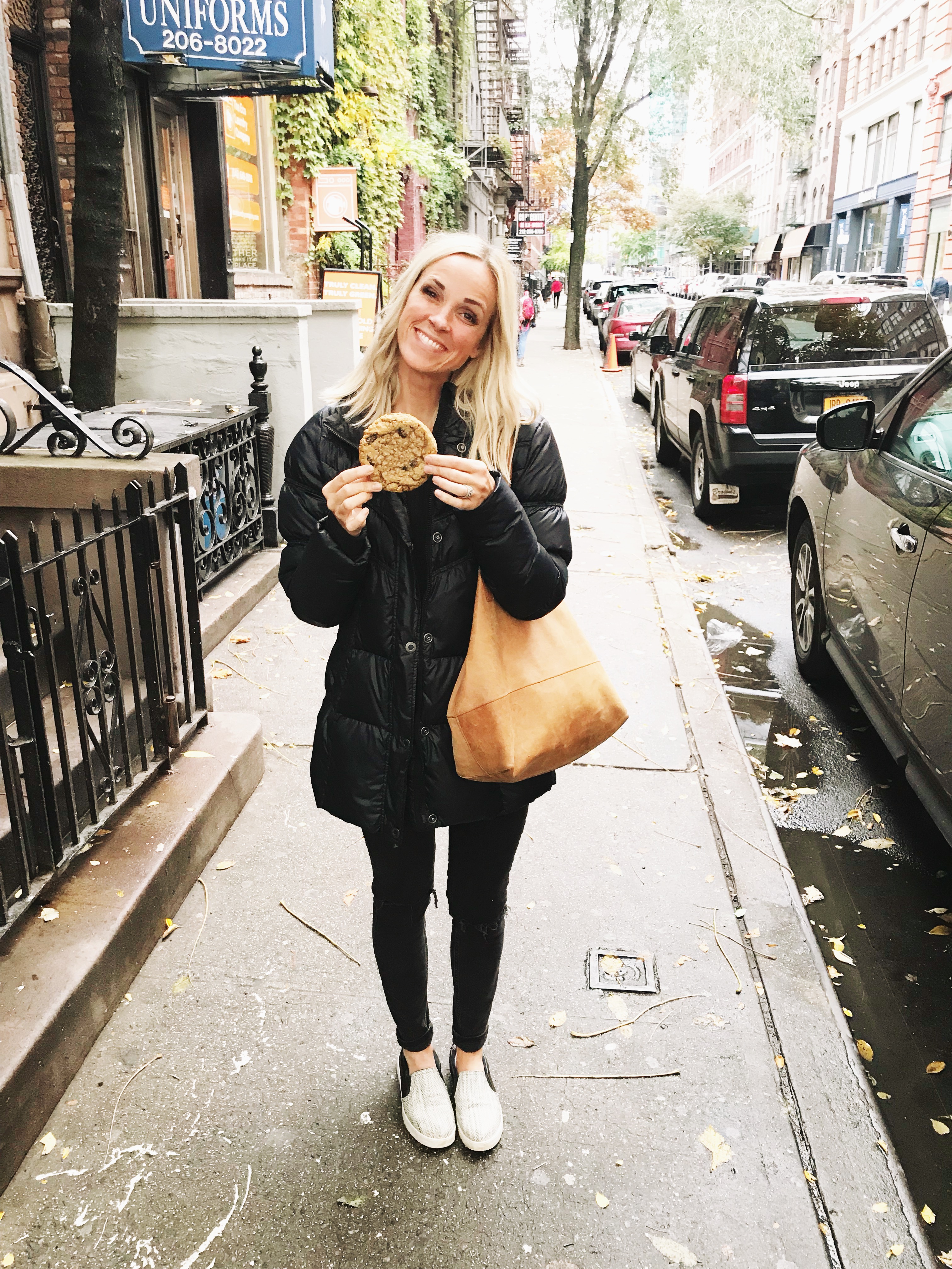 A woman in a black jacket stands on a city sidewalk, smiling and holding a large cookie—perhaps thinking about macro counting as she enjoys her treat. She has a tan bag over her shoulder and wears black pants and white shoes. Cars and trees line the street.