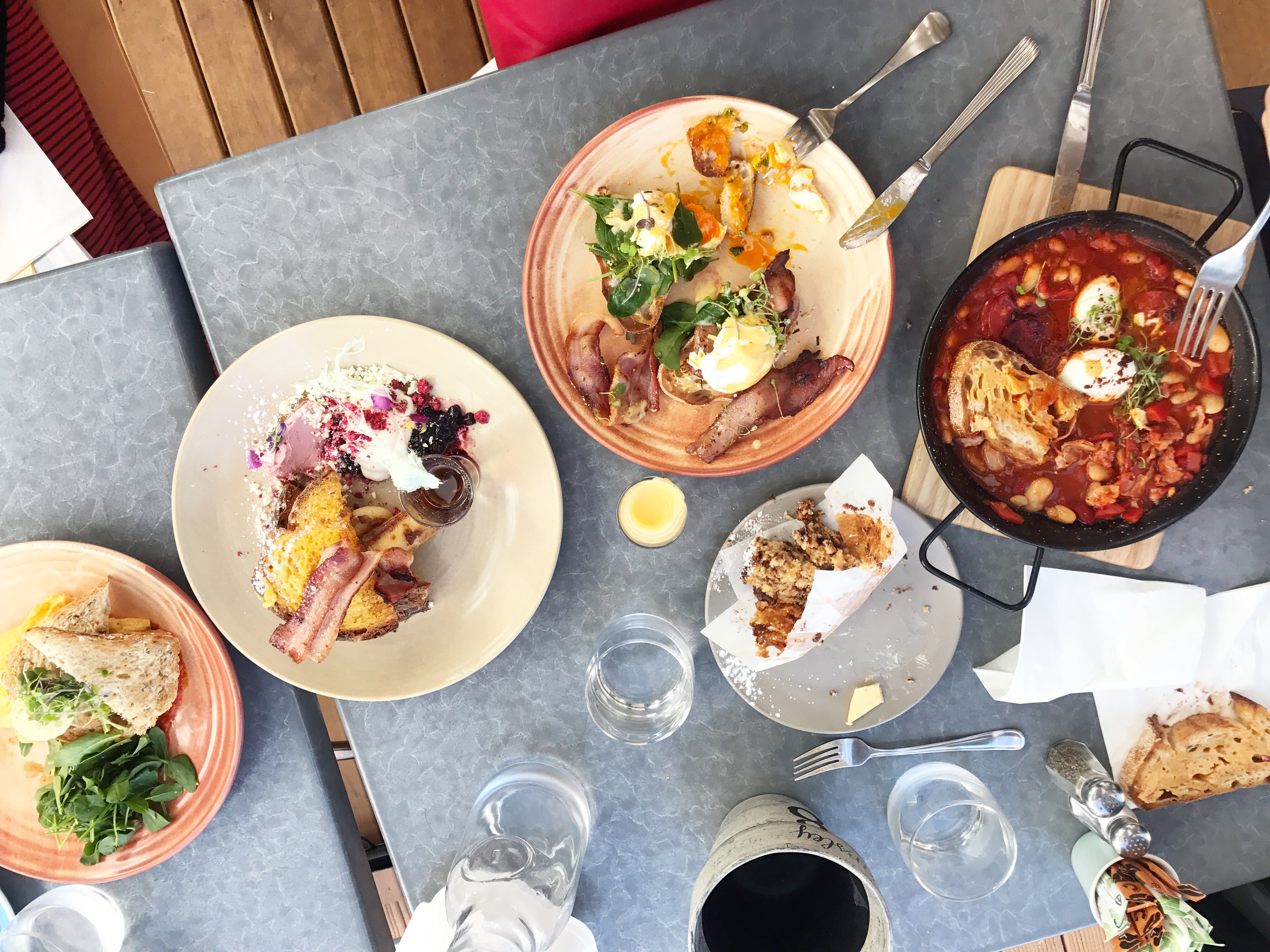 Top-down view of a table with various brunch dishes, including poached eggs on toast, bacon, grilled vegetables, bread, and a skillet with tomato-based stew—perfect for anyone interested in macro counting. Plates and glasses are partially filled on a gray tabletop.