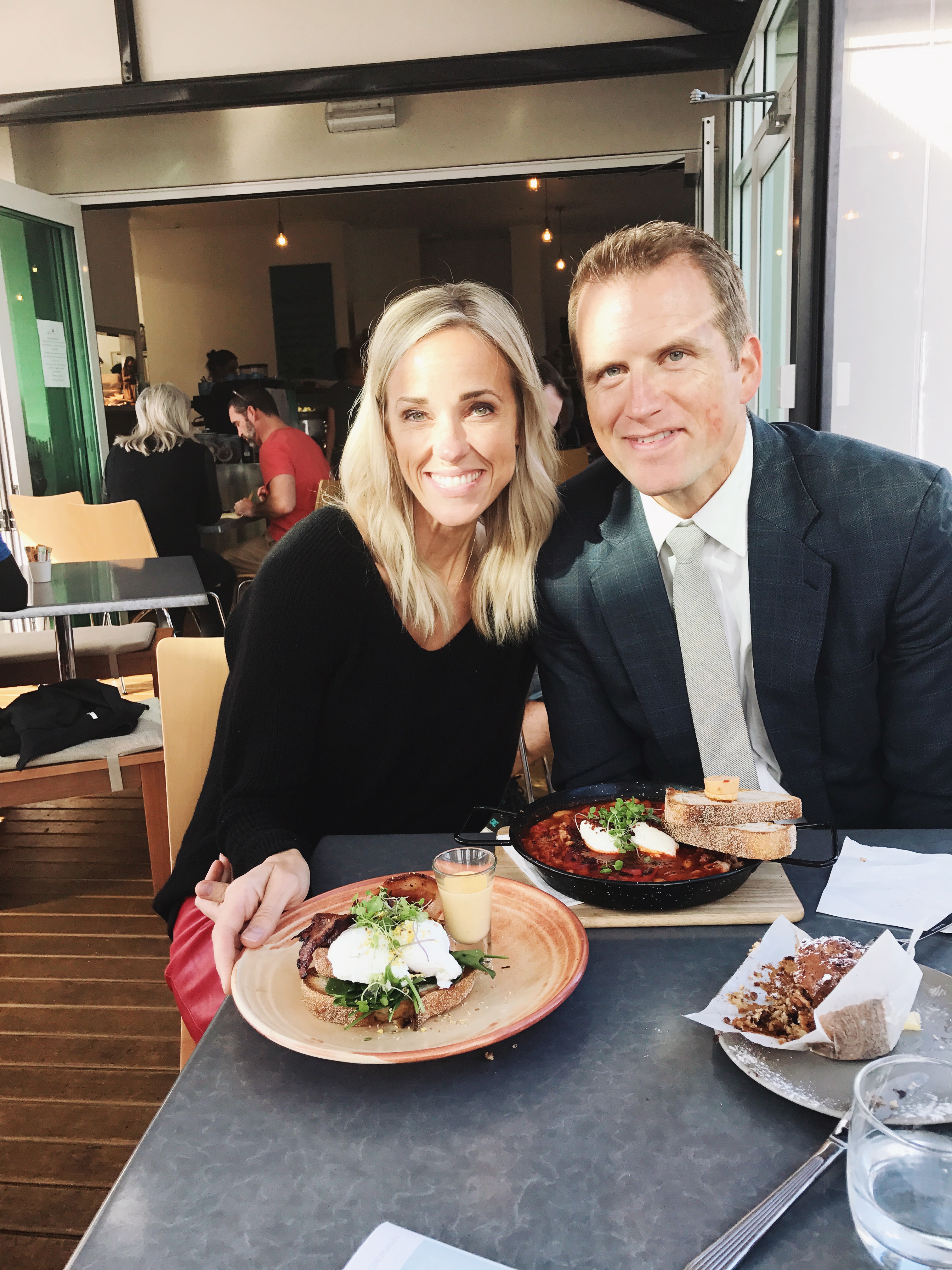 A smiling woman and man sit at a restaurant table with plates of food and drinks, thoughtfully macro counting as they pose for the photo. Sunlight streams in through large windows behind them.