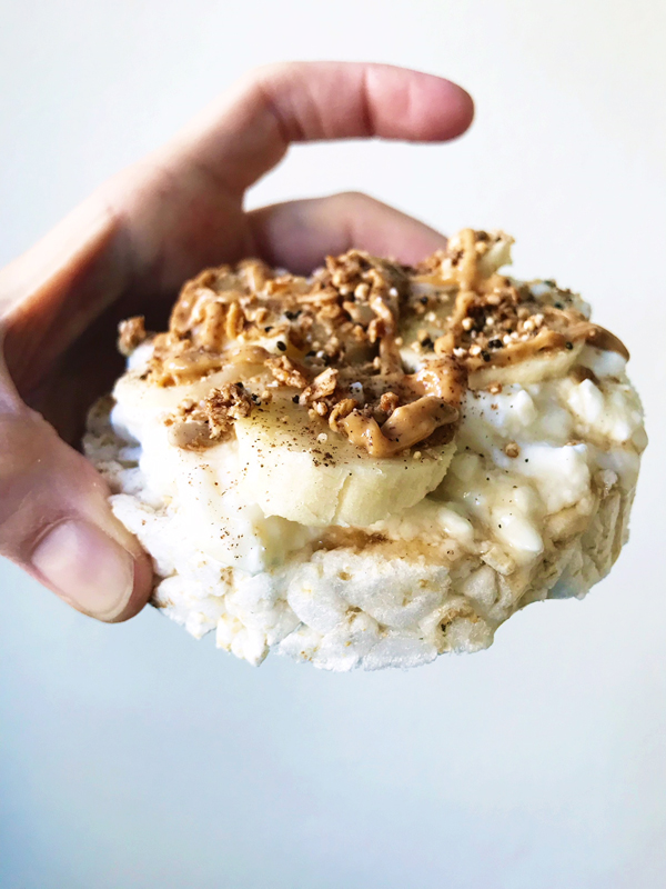 A hand holds a rice cake topped with banana slices, peanut butter, and sprinkled granola against a plain background—a delicious snack idea for a day of eating.