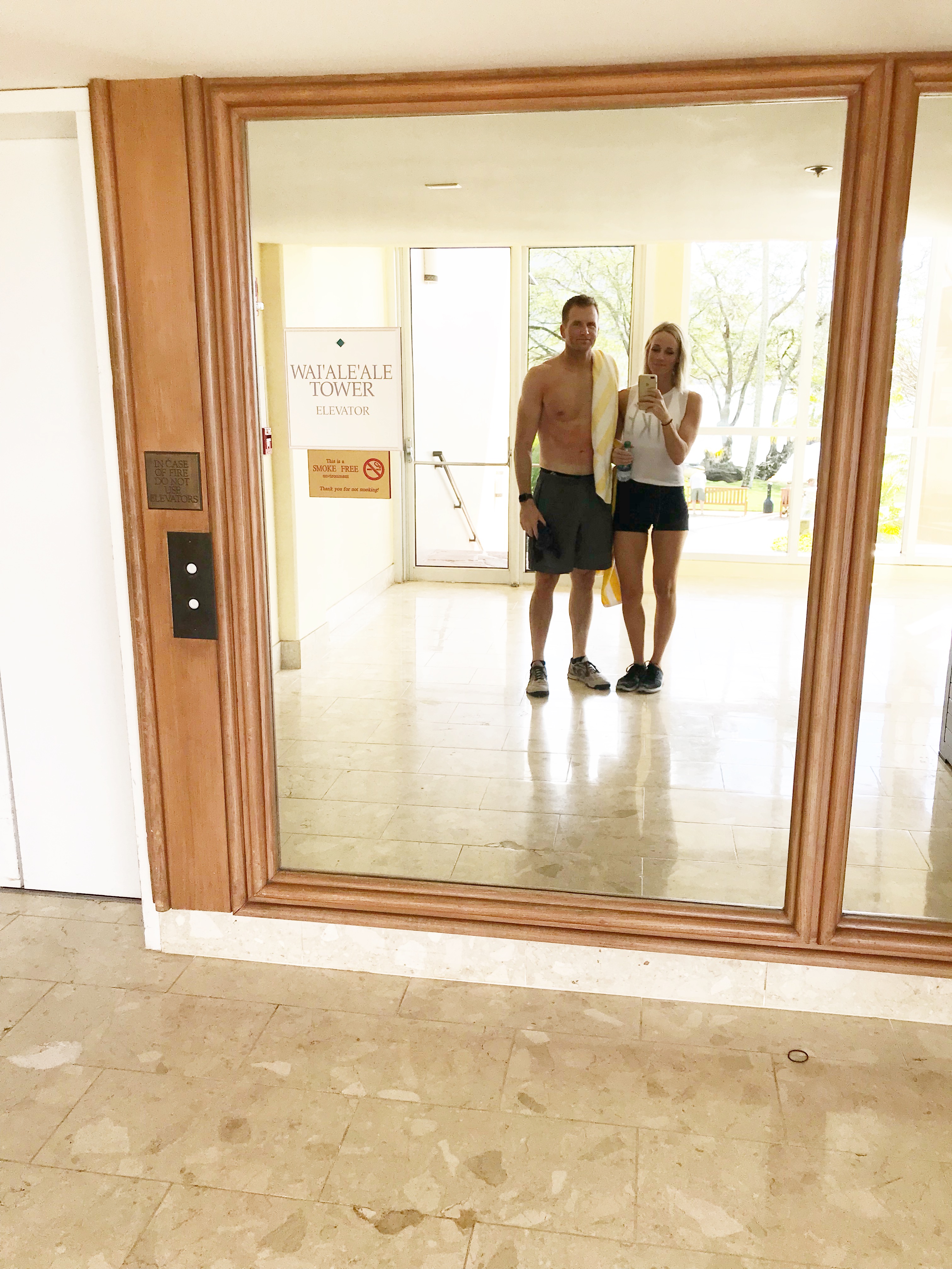 A man and a woman stand side by side, reflected in a large wall mirror in a bright lobby. The man is shirtless with a towel over his shoulder, perhaps after an intense workout focused on macro counting; the woman holds a phone for a selfie.