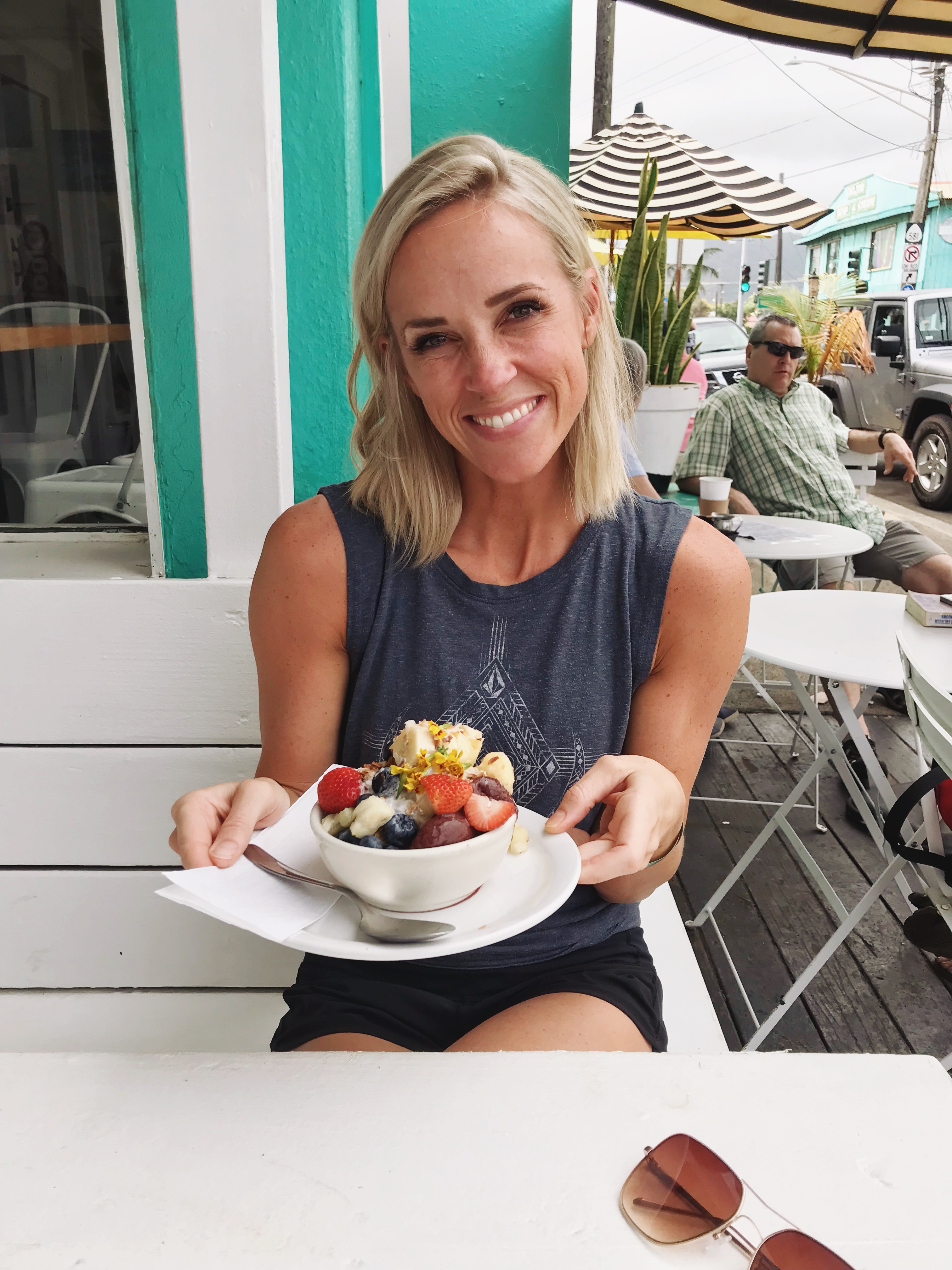 A woman with blonde hair smiles while holding a bowl of fruit and ice cream at an outdoor cafe, thoughtfully enjoying her treat while macro counting. Sunglasses rest on the white table in front of her. Other diners and colorful buildings are visible in the background.