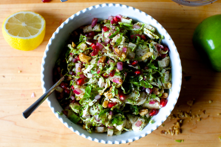 A bowl of colorful salad with chopped greens, red onions, pomegranate seeds, and nuts, with a spoon inside—perfect for brightening up your Thanksgiving menu. A lemon half and a green apple are on the wooden table beside the bowl.