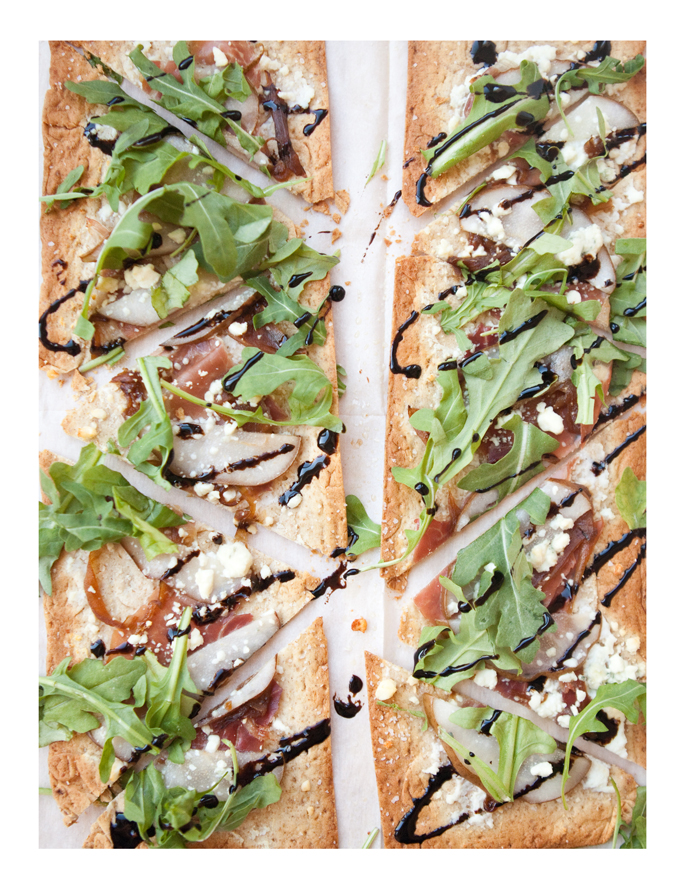 Flatbread topped with arugula, sliced pears, crumbled cheese, and a drizzle of balsamic glaze, cut into rectangular pieces and arranged on parchment paper—a delicious addition to any Thanksgiving menu.