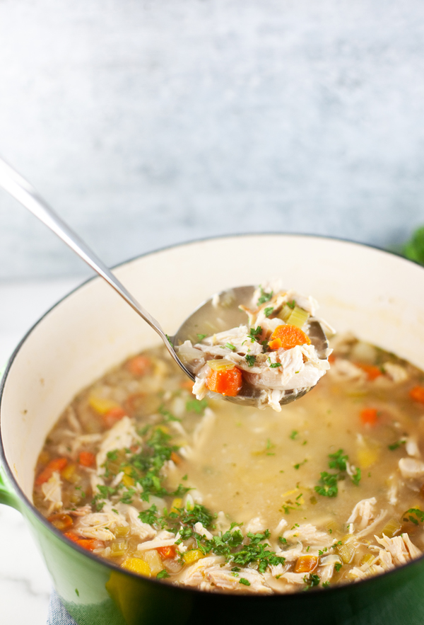 A ladle lifts homemade lemony chicken soup with carrots, celery, and shredded chicken from a green pot, garnished with fresh herbs.