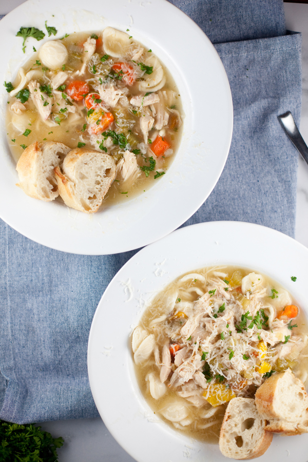 Two white bowls of lemony chicken soup with noodles and vegetables, topped with shredded cheese and chopped herbs, served with slices of crusty bread on a blue cloth.