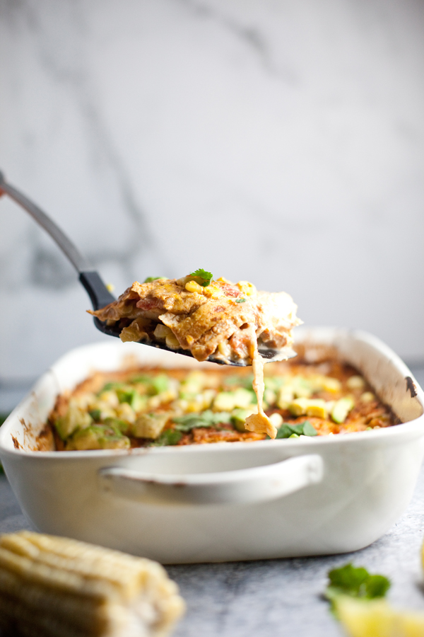 A close-up of a baking dish with a cheesy, baked Green Chili Chicken Bake casserole. A spatula lifts a serving, showing melted cheese and layers inside. The casserole is topped with herbs and sits on a gray surface. Corn and a lime wedge are in the foreground.