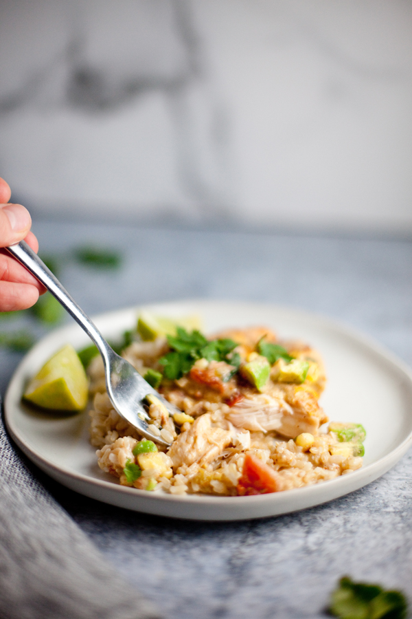 A hand holding a fork digs into a plate of Green Chili Chicken Bake, nestled alongside rice and vegetables, garnished with cilantro, with a lime wedge on the side.