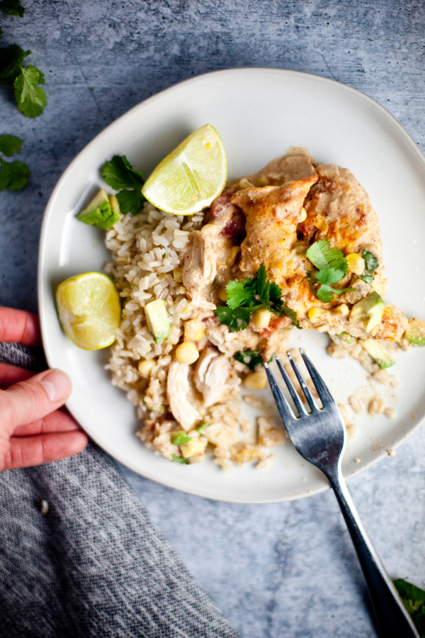 A plate of Green Chili Chicken Bake with brown rice, corn, and cilantro, garnished with lime wedges. A hand holds the plate, and a fork rests on it.