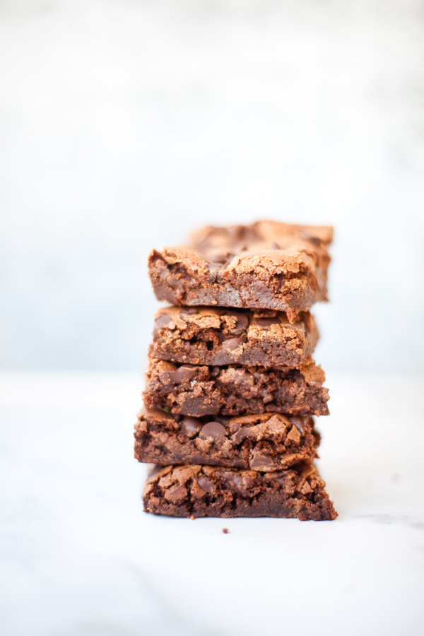 A stack of four chocolate brownies—perhaps the best brownies ever—sits on a white surface with a light, blurred background. The brownies look moist with visible chocolate chunks.