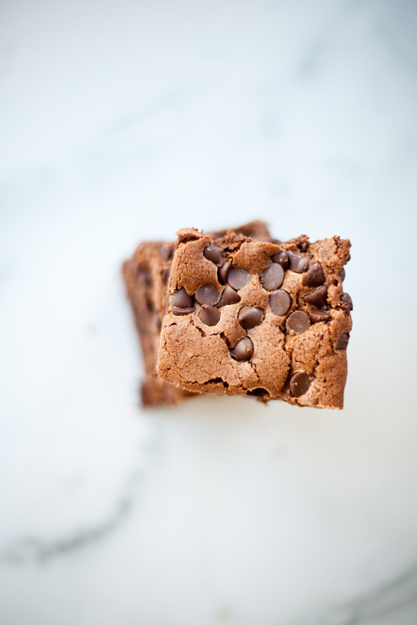Two of the best brownies ever, filled with chocolate chips, are stacked on a white marble surface and viewed from above.