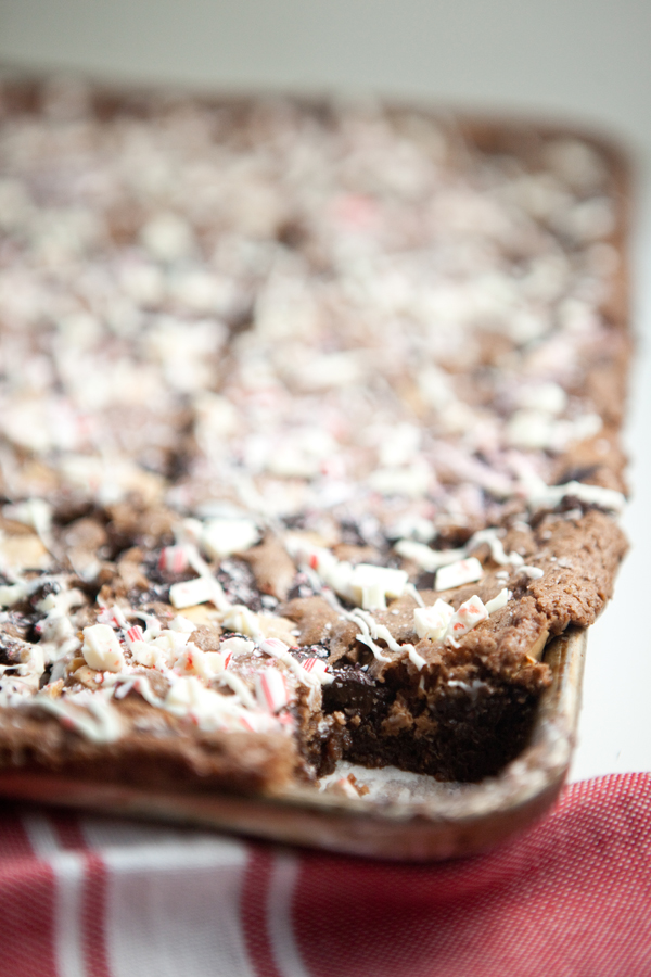 A close-up of a large, rectangular chocolate brownie—quite possibly the best brownies ever—topped with crushed peppermint candies and white drizzle, with one piece missing from the corner.