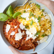 A bowl with Instant Pot Pork Ragu—shredded meat in tomato sauce—spaghetti squash, fresh basil, and shaved parmesan cheese, held by two hands with a fork.
