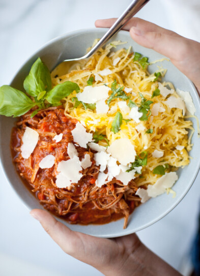 A bowl with Instant Pot Pork Ragu—shredded meat in tomato sauce—spaghetti squash, fresh basil, and shaved parmesan cheese, held by two hands with a fork.