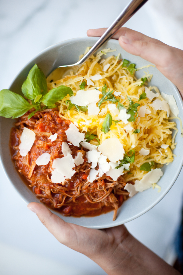 A bowl with Instant Pot Pork Ragu—shredded meat in tomato sauce—spaghetti squash, fresh basil, and shaved parmesan cheese, held by two hands with a fork.