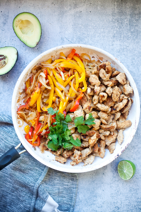 A skillet with cooked sliced fajita chicken and veggies—sautéed yellow and red bell peppers, onions, and fresh cilantro. Half an avocado, half a lime, and a towel sit beside the pan on a gray surface.