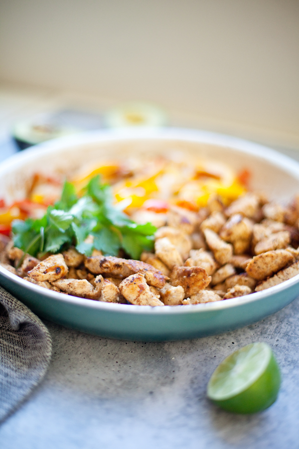 A bowl of cooked fajita chicken and veggies with sliced bell peppers and fresh cilantro. A lime wedge sits nearby on the table.