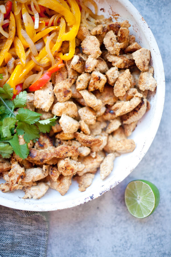 A plate with seasoned cooked chicken pieces, sautéed yellow and red bell peppers, fresh cilantro, and a halved lime on the side—this vibrant dish perfectly showcases classic fajita chicken and veggies, set on a light gray surface.