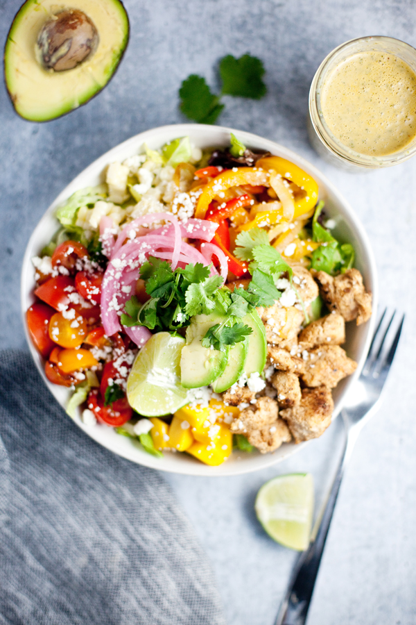 A colorful salad bowl featuring fajita chicken and veggies, creamy avocado, pickled onions, tomatoes, peppers, cilantro, lime wedges, and cheese. A glass of dressing, a fork, and a halved avocado are arranged beside the bowl on a gray table.