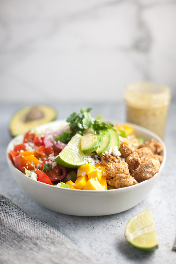 A bowl of salad with fajita chicken and veggies, avocado, cherry tomatoes, bell peppers, cilantro, and a lime wedge. A halved avocado, a jar of dressing, and a lime wedge are in the background.