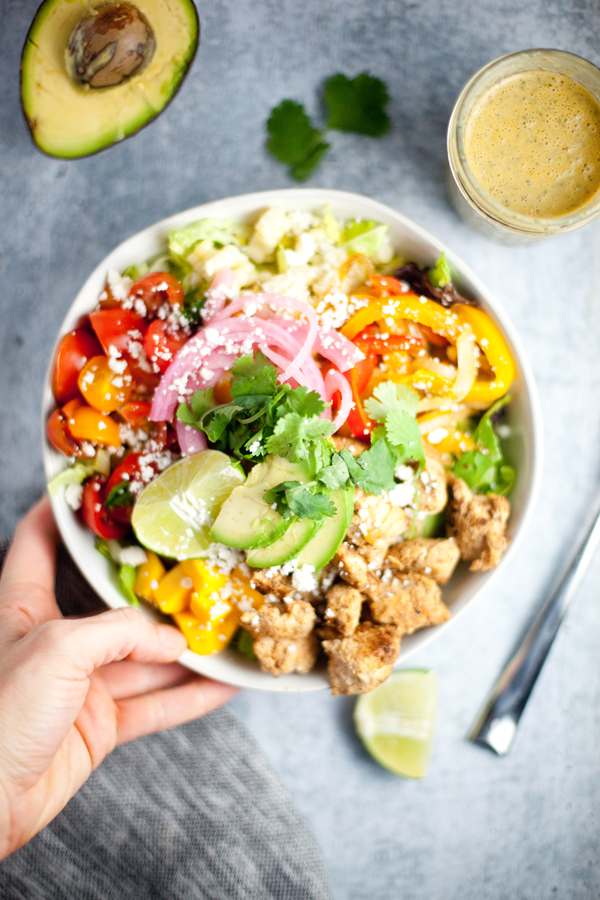 A hand holds a bowl filled with colorful fajita chicken and veggies, creamy avocado slices, pickled onions, lime wedges, cilantro, and chunks of cooked chicken. An avocado half, lime wedge, fork, and glass of dressing are nearby.