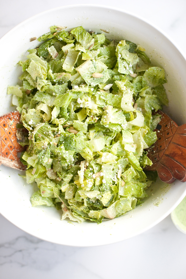 A white bowl filled with chopped romaine lettuce, grated cheese, and Caesar dressing, being tossed with wooden salad servers on a marble surface—a perfect example of a Light Mexican Caesar salad.