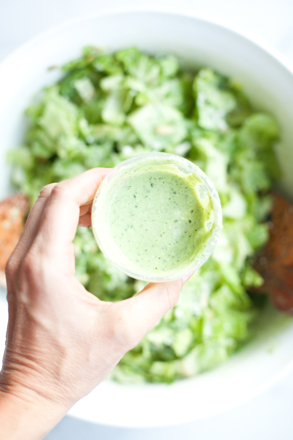 A hand holds a small container of creamy green Light Mexican Caesar salad dressing above a bowl of leafy green salad.