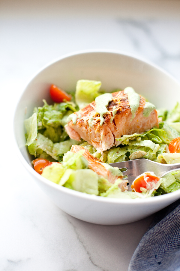 A white bowl filled with salad greens, cherry tomatoes, and a piece of grilled salmon topped with creamy dressing, reminiscent of a Light Mexican Caesar salad, with a fork resting inside the bowl.