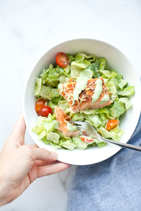 A hand holds a bowl of Light Mexican Caesar salad with lettuce, cherry tomatoes, grilled salmon, and green dressing. A fork rests in the bowl.