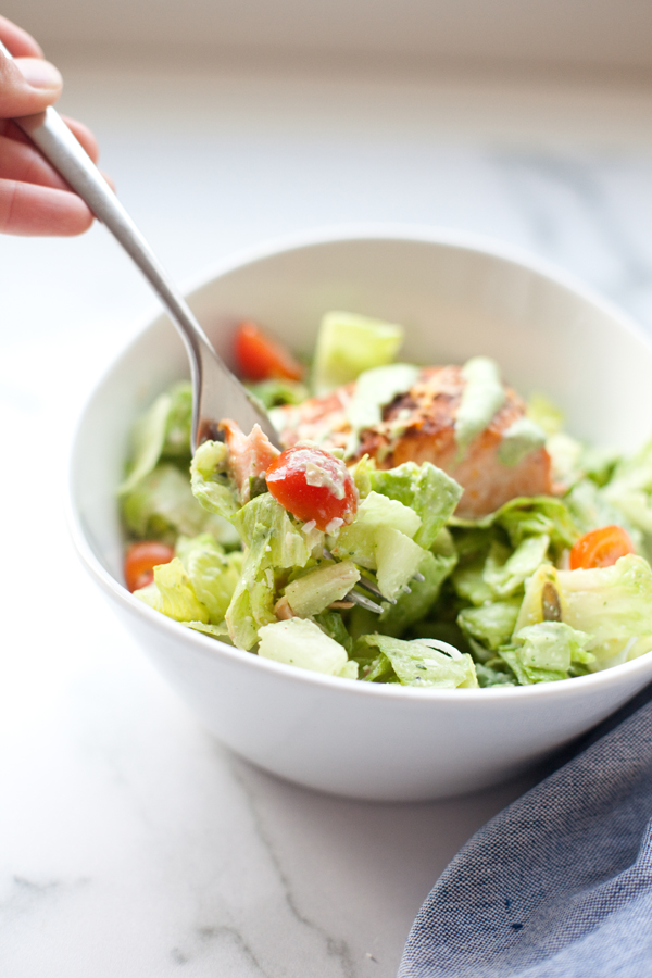 A hand holding a fork over a bowl of fresh Light Mexican Caesar salad with lettuce, cherry tomatoes, cucumbers, and grilled chicken on a marble surface.
