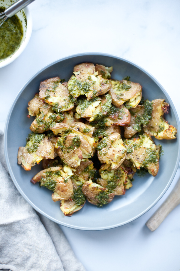 A blue plate filled with smashed potatoes topped with vibrant green herb sauce, set on a white surface next to a small bowl of sauce and a light-colored napkin.